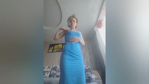 A video of a woman with short blonde hair in a blue dress, standing in a bedroom with a floral-patterned bedspread and modern decor.