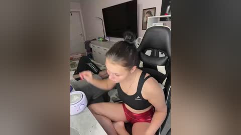 Video of a young woman with dark hair tied back, wearing a black sports bra and red shorts, playing a video game on a white desk in a cluttered, dimly lit room with a black gaming chair.