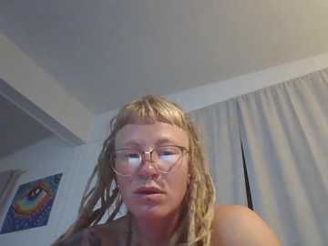 A video of a young woman with light skin, long blonde dreadlocks, and glasses, wearing a sleeveless top. She is indoors, with a white ceiling and a partially visible curtain.