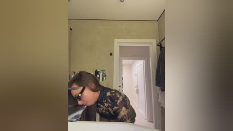 A video of a woman with glasses and long brown hair in a bathroom, leaning over a counter, looking into a mirror, beige walls, and towels hanging on a rack in the background.