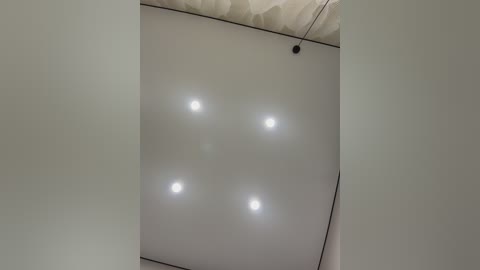 Video of a minimalist ceiling with four evenly spaced circular recessed lights, surrounded by soft, diffused light, and a single, thin black cord hanging down.
