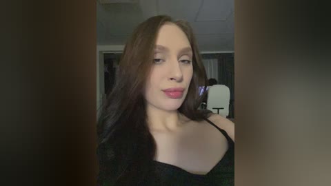 Video of a fair-skinned woman with long, straight brown hair, wearing a black off-shoulder top, standing in a dimly lit room with white ceiling tiles and dark furniture in the background.