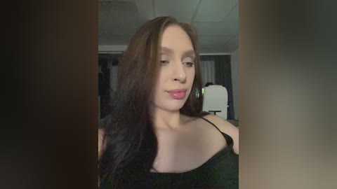 Video of a fair-skinned woman with long, dark brown hair, wearing a black off-shoulder top, in an indoor setting with dim lighting and indistinct background.