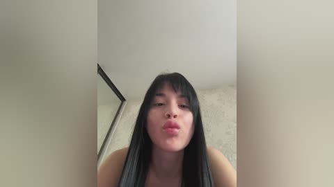 A video of an Asian woman with straight black hair, puckered lips, and fair skin, captured from a low angle. The background shows a plain, off-white wall with a slight texture and a black trim.