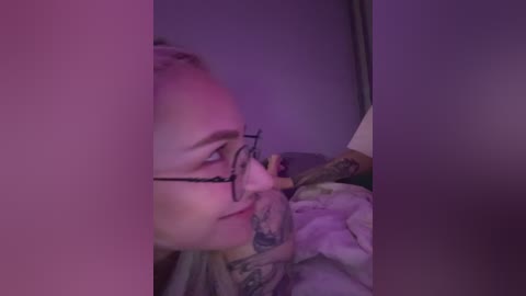 Video of a tattooed, blonde woman wearing glasses, partially obscured by a pinkish-purple curtain. She is lying on a bed with a white sheet, her face in profile, looking down.