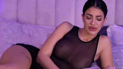Video of a light-skinned woman with dark hair, wearing a see-through black dress, lying on a bed with white sheets, in a softly lit room.