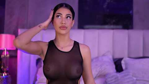 Video of a woman with long dark hair, light skin, and large breasts, wearing a sheer black top, in a modern bedroom with a white bed and purple lighting.