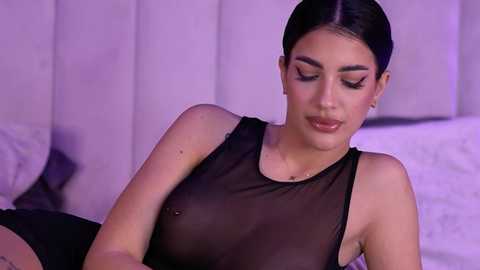 Video of a Latina woman with olive skin, dark hair tied back, wearing a sheer black top that reveals her nipples, lying on a bed with white sheets, in a dimly lit room.