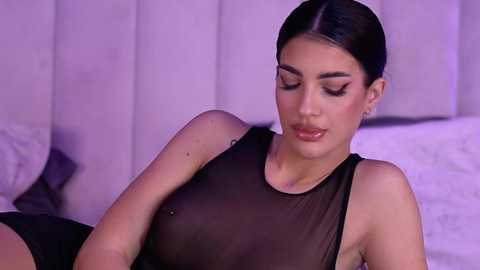 Video of a young woman with medium skin tone, dark hair tied back, wearing a sheer black top revealing her small breasts, lying on a bed with white pillows and a headboard.