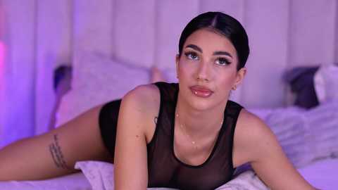 Video of a young Latina woman with dark hair, wearing a sheer black top and black underwear, lying on a bed with white pillows and a white headboard.