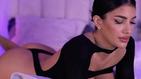 Video of a light-skinned woman with dark hair, wearing a black long-sleeved top and thong, lying on a bed with white sheets, under soft purple lighting.