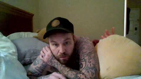 Video of a bearded, tattooed man in a black cap and pink shirt, lying on a bed with yellow and green pillows, in a dimly lit room.