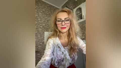 Video of a smiling, fair-skinned woman with long, wavy blonde hair, wearing glasses and a white lace blouse, standing in a rustic room with a brick wall and white-framed window.