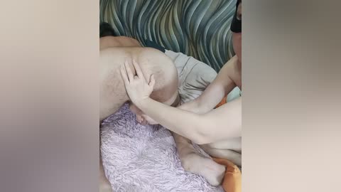 A video captures a close-up of a man and woman in an intimate moment. The woman, with fair skin and a slender build, is bent over a bed, her buttocks exposed. The man, with a darker complexion, is seen pressing his hand against her lower back. The background includes a striped bedspread and a fluffy, pink blanket.