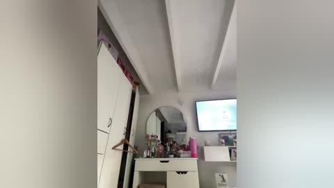 Video of a minimalist, modern bedroom with white walls and exposed beams, featuring a vanity table with makeup items, a mirror, and a TV displaying a website.