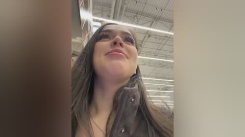 Video of a young woman with long, straight brown hair, wearing a brown jacket, smiling upward, taken indoors with a metallic ceiling and fluorescent lights.