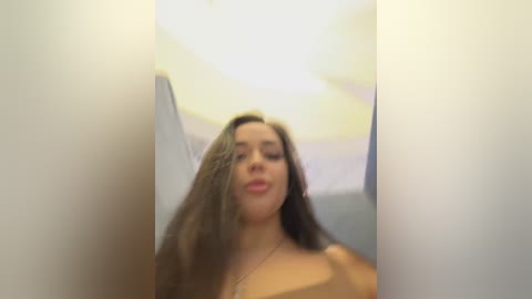 Video of a young woman with long, wavy brown hair, light skin, and a slender physique, smiling, taken from a low angle, with blurred background.