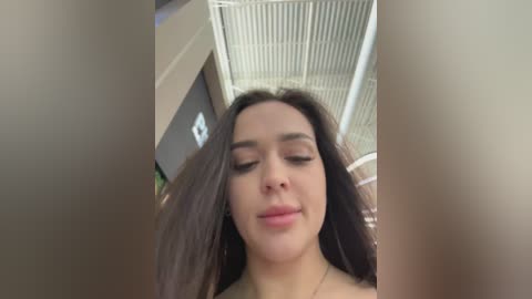 Video of a young woman with long, dark hair, fair skin, and closed eyes, wearing a black top, standing in an indoor room with beige walls and a white ceiling.