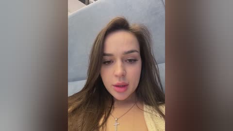 Video of a young woman with fair skin, long brown hair, and a neutral expression. She wears a white top and a cross necklace. Background is a light blue sofa.