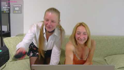 Video of two women, one blonde in a white shirt, the other with long hair in an orange top, playing a game on a laptop on a green sofa.