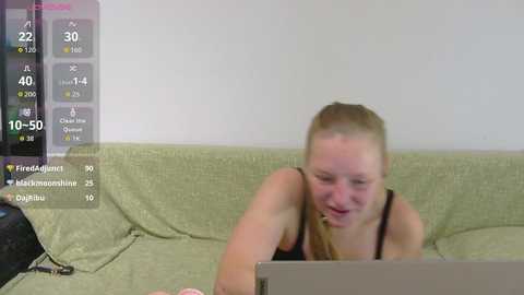 A video of a young, fair-skinned woman with long blonde hair in a black tank top, sitting on a green couch, focused on a laptop, with a digital overlay showing streaming stats.