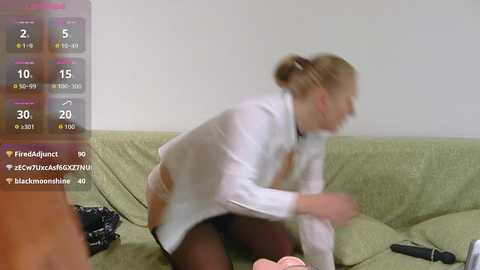 Video of a blonde woman in a white shirt and black panties, crouched on a green couch, surrounded by video game controllers.