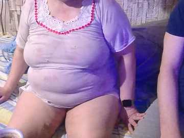Video of a chubby, light-skinned woman wearing a wet, see-through white top revealing her large breasts and nipples. She sits on a bed with a man's arm visible. Background includes a beige wall and a blue blanket.