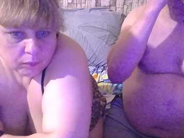 Video of two overweight, fair-skinned women with short hair, one partially clothed, lying on a bed with a leopard-print bra. Background includes a messy bed and a wall with a patterned wallpaper.