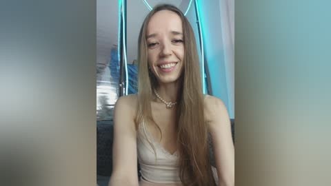 Video of a young, smiling Caucasian woman with long, straight brown hair, wearing a light beige spaghetti-strap top and pearl necklace, seated indoors against a modern, teal-lit backdrop.