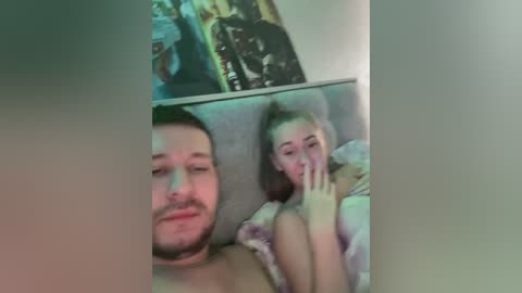 Video of a bearded man and a blonde woman, both topless, lying in bed with a blurred background, suggesting a casual, intimate moment.