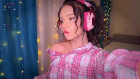 Video of a young woman with curly brown hair, wearing pink headphones, plaid shirt, and a pensive expression. Background includes string lights and a bed.