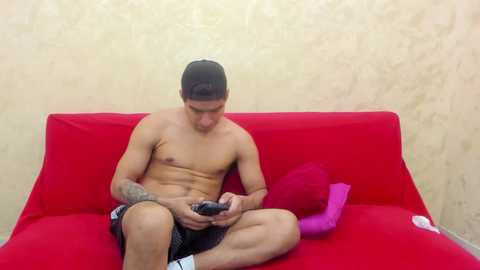 Video of a shirtless, muscular, light-skinned man with short dark hair, sitting on a bright red sofa against a beige textured wall. He's focused on a smartphone, wearing black shorts and white socks.