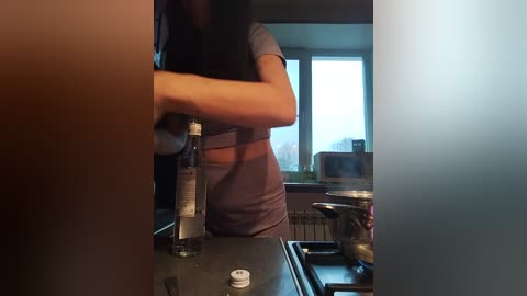 A video of a person with medium brown skin, long black hair, and wearing a gray shirt, pouring olive oil into a pan on a stove. The background shows a modern kitchen with a window and a radiator.