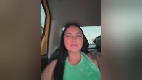 A video of a Latina woman with long black hair, wearing a green tank top, sitting in a car, blurred background.