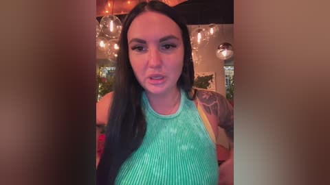 Video of a young woman with long dark hair, wearing a green ribbed tank top, standing indoors with chandeliers, green plants, and a bar in the background.