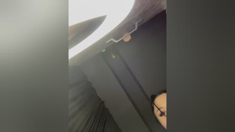 Video of a person with glasses partially visible, wearing a black top, and standing in a dimly lit room with a white ceiling light and a wooden ceiling beam.