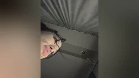 Video of a person's face reflected in a mirror, with a light pink complexion, short dark hair, and closed eyes. The background shows a grey pillow and a wooden door knob.