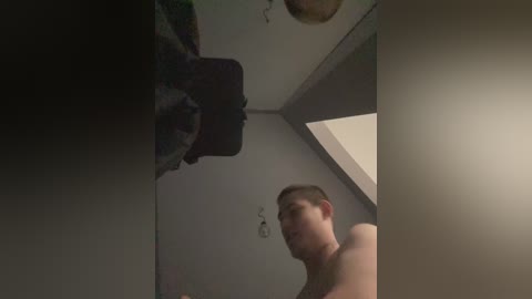 A dimly lit video captures a shirtless, pale-skinned man with short brown hair standing near a mirror in a small, sparsely decorated room. The room's walls are white, and a light fixture is visible on the ceiling.