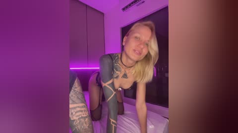 Video of a blonde woman with tattoos, wearing a revealing black bodysuit, on all fours in a dimly lit room with purple lighting.