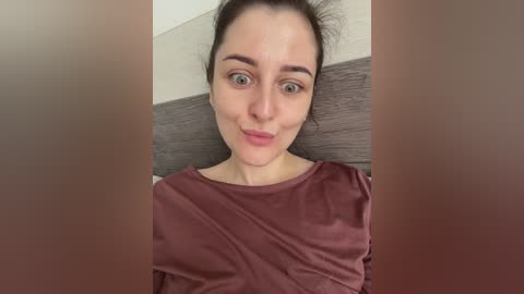 Video of a young woman with fair skin, dark hair tied back, wearing a maroon shirt, sitting against a grey headboard, smiling softly.