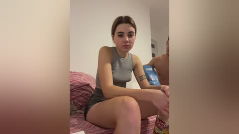 Video of a young woman with light skin and brown hair, sitting on a bed with a pink and white striped blanket, wearing a grey tank top and shorts, holding a blue water bottle, in a dimly lit room.