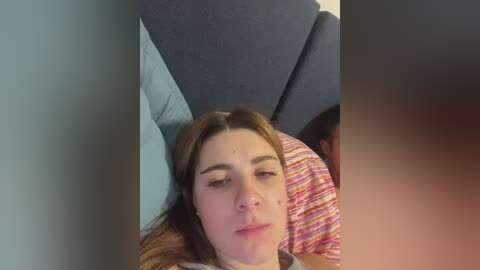 Video of a young woman with fair skin, brown hair, and light makeup lying on a gray couch. She has a neutral expression. The background includes a striped pillow and a gray cushion.