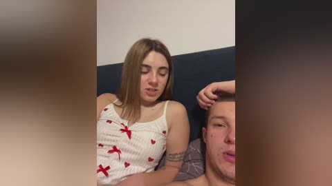 Video of a young Caucasian woman with straight, shoulder-length brown hair, wearing a white tank top with red heart patterns, lying on a bed next to a man. The background is a dark blue headboard against a white wall.