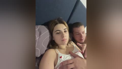 Video of a young woman with light skin, wearing a white tank top, lying on a bed with a dark blue headboard, holding a baby, while a man with short brown hair and light skin, wearing a white shirt, lies beside her.