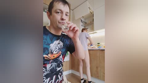 Video of a man in a dark T-shirt featuring a cartoon duck, holding a glass in a modern kitchen; another person in the background.