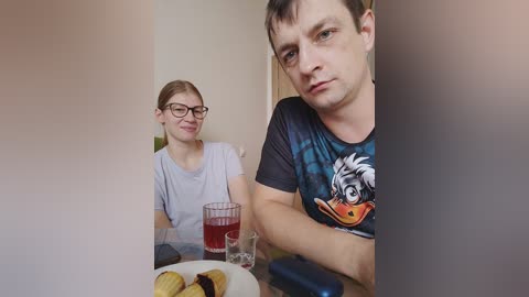 Video of a young woman with glasses and a light blue shirt, seated beside a man in a blue shirt with a duck graphic, eating cookies at a table.