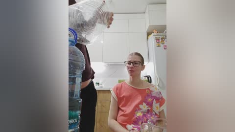 Video of a woman with glasses and light skin, wearing a coral shirt with floral pattern, sitting at a kitchen table, pouring water from a plastic bottle into a glass.