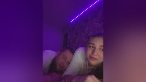 Video of a young woman with long dark hair and a man with a beard, both lying in bed with purple LED lights.