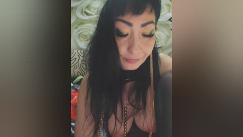 Video of a woman with long, straight black hair and bangs, wearing heavy makeup and a black choker necklace, against a background of beige and white floral wallpaper.