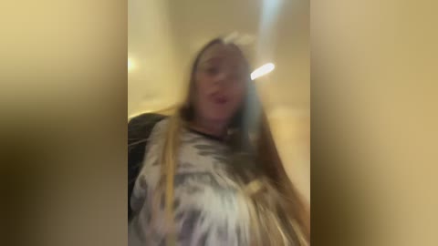 A blurry video of a woman with long, light blonde hair, wearing a white and black patterned robe, standing in a dimly lit, beige hallway. The image is slightly out of focus.